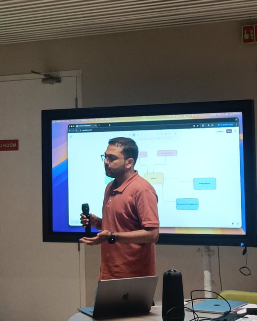 A man in a terracotta polo shirt is giving a presentation, standing in front of a large screen displaying a flowchart while holding a microphone.