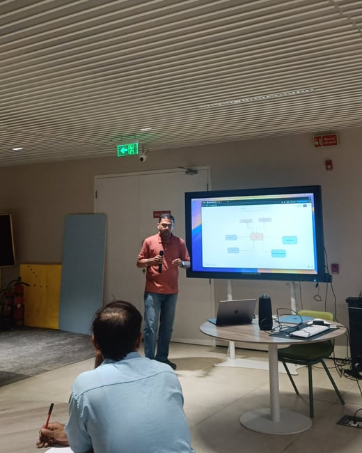A man in a red polo shirt is giving a presentation in a modern office, standing next to a large screen displaying a diagram while an audience member listens in the foreground.