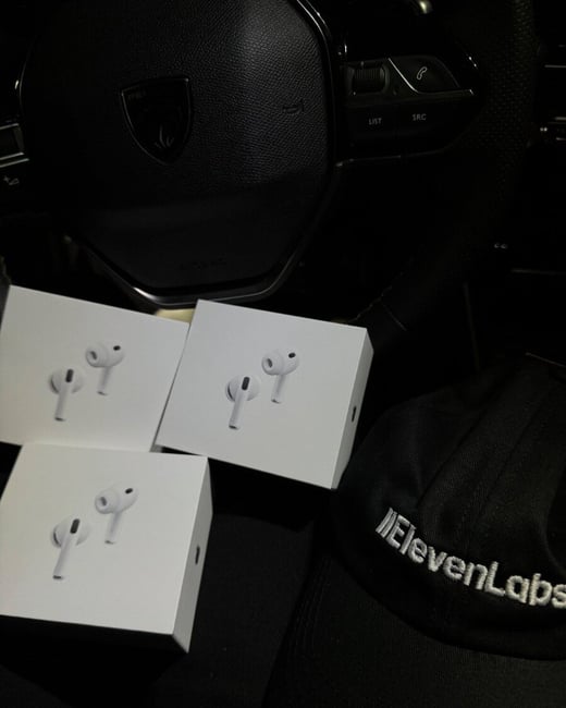 A low-light photograph showing three white boxes of Apple AirPods resting near a black baseball cap embroidered with the word "ElevenLabs," all positioned near the steering wheel of a car.