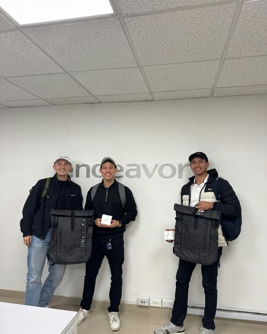 A photo of three young men standing indoors in an office environment, posing with black backpacks labeled '11ElevenLab' and small white boxes, in front of a wall featuring the company name 'endeavor'.