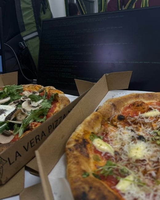A close-up photograph captures two gourmet pizzas in open cardboard boxes resting on a desk, with a dark computer monitor displaying lines of code visible in the background.