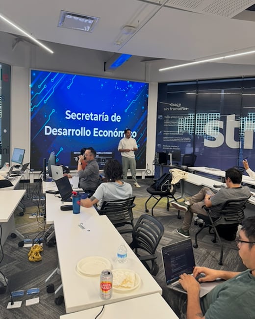 A man is giving a presentation in a modern office setting to a group of people seated at desks with their laptops open.