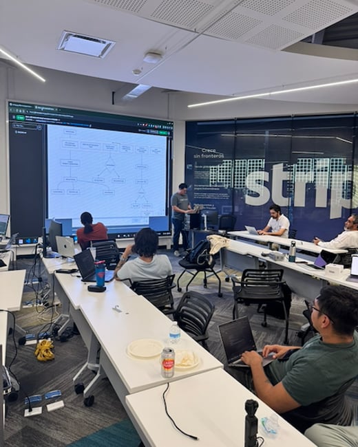 A group of people in a modern office environment attending a presentation or workshop, with a large screen displaying a technical flowchart and 'Stripe' branding on the wall.