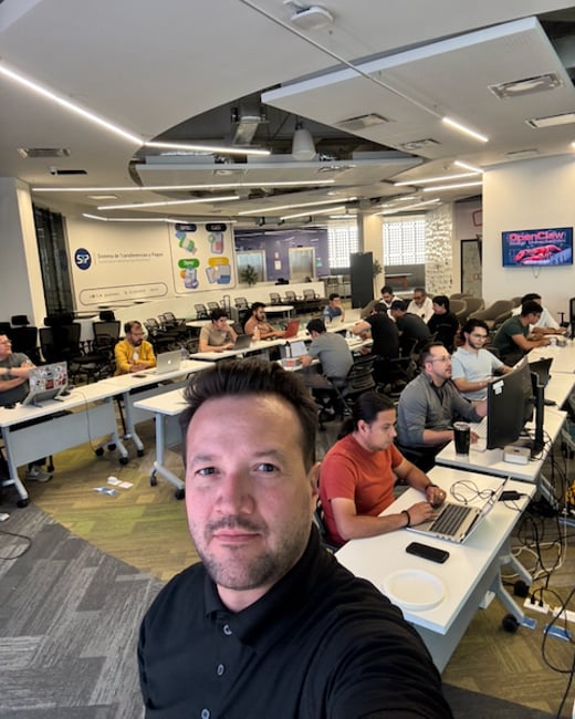 A man takes a selfie in a modern, open-plan office where several people are working on laptops at long white tables.