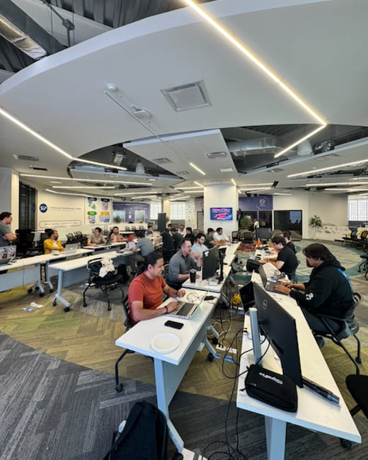 A group of people are working on laptops in a spacious, modern office with unique curved ceiling panels and long white communal desks.