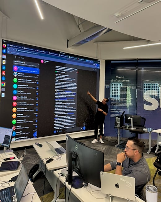 A man stands in a modern office pointing at a large digital screen displaying a messaging application, while another man works at a desk in the foreground.