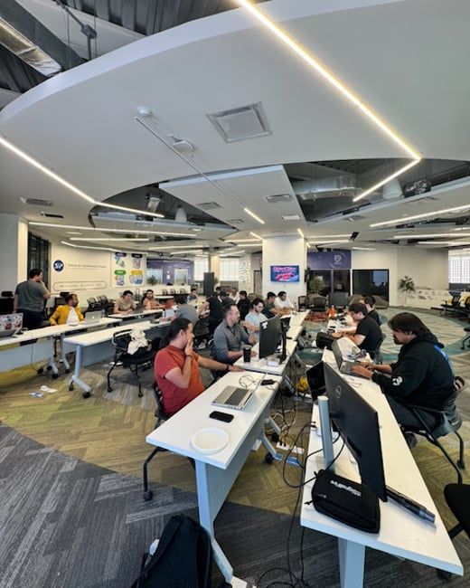 A wide-angle photograph of a modern, open-plan office where a group of people are working at long white desks equipped with laptops and monitors.