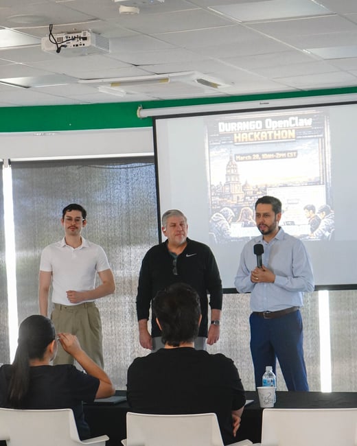 A man in a light blue shirt speaks into a microphone during a hackathon presentation, while two other men stand beside him and an audience sits in the foreground.