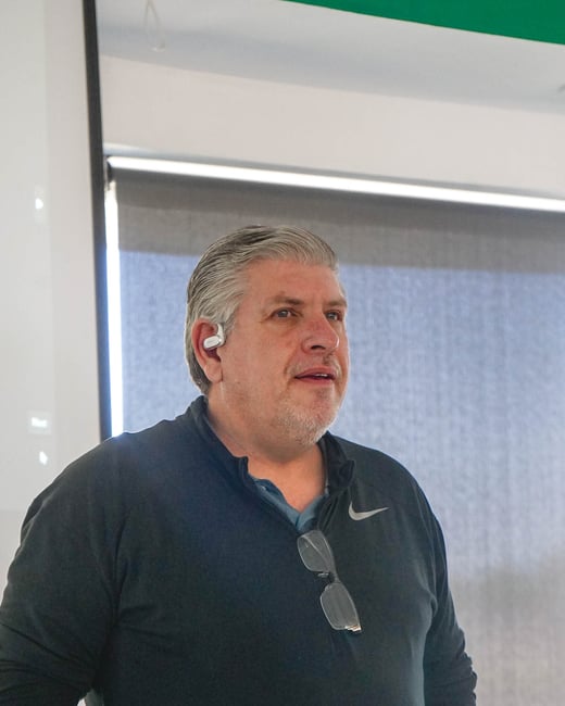 A medium shot of a middle-aged man with grey hair standing indoors, wearing a black long-sleeve Nike shirt and a white earbud.