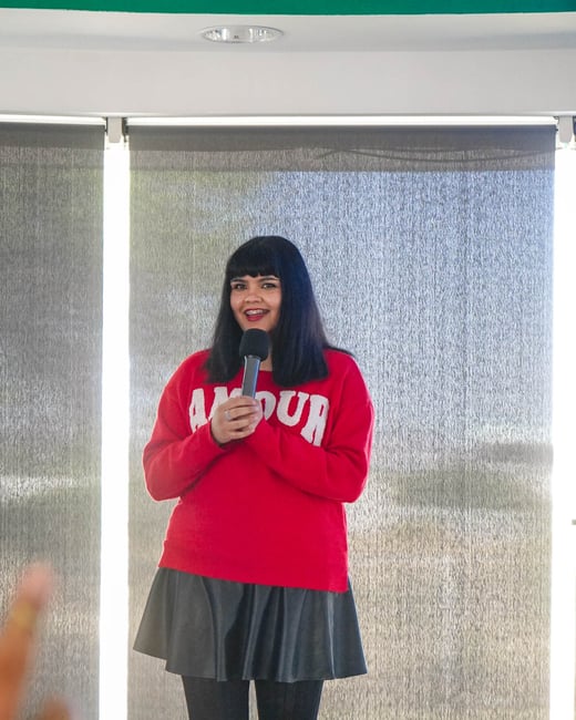 A woman with dark hair and bangs is standing indoors, holding a microphone and wearing a red sweater with the word 'AMOUR' on it.