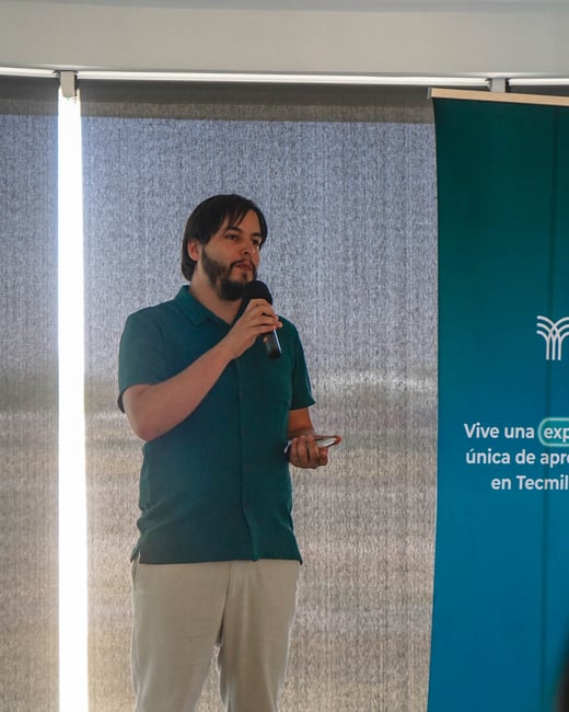 A man with a beard is standing in a room, speaking into a microphone while holding a phone, positioned next to a blue promotional banner.