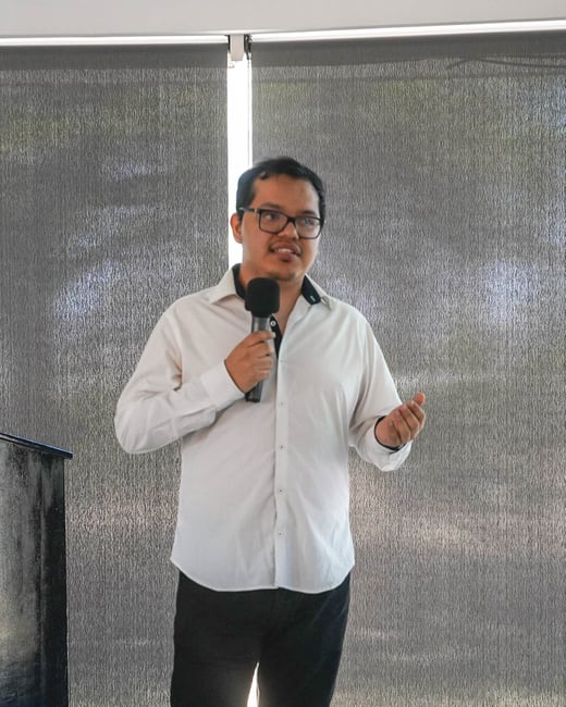 A man in a white shirt and black pants stands indoors, speaking into a microphone.
