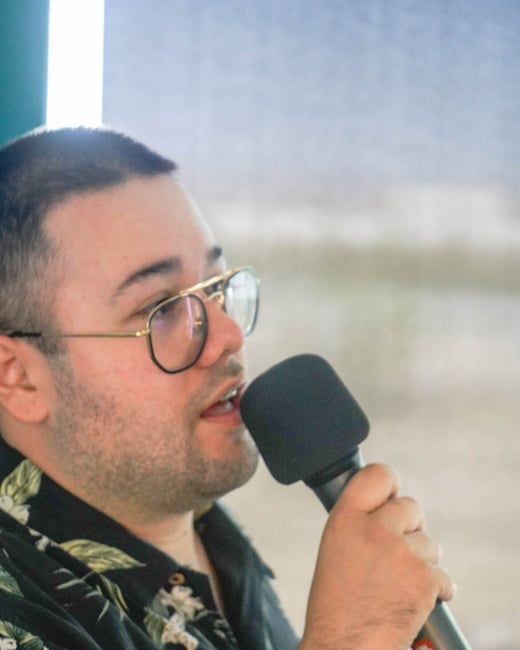 A man with glasses and a floral shirt is speaking into a microphone.