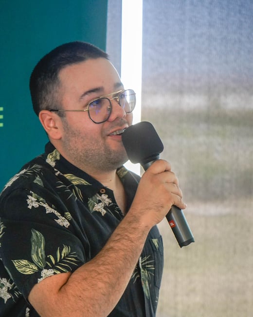 A man wearing glasses and a floral shirt is speaking into a microphone during an indoor event.