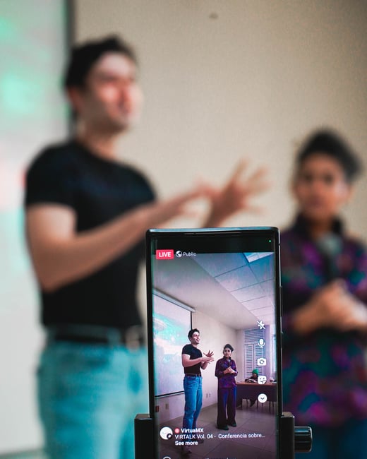 A smartphone mounted on a tripod is live-streaming a presentation given by two people in an indoor setting.