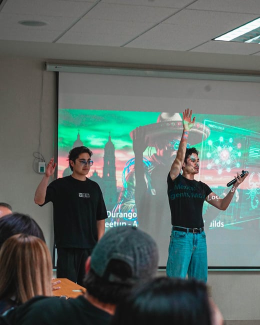 Two young men are giving a presentation in front of a projector screen in a classroom or meeting room, with an audience visible in the foreground.