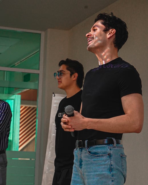 A man in a black t-shirt and blue jeans holds a microphone and smiles while standing next to another man at an indoor event.
