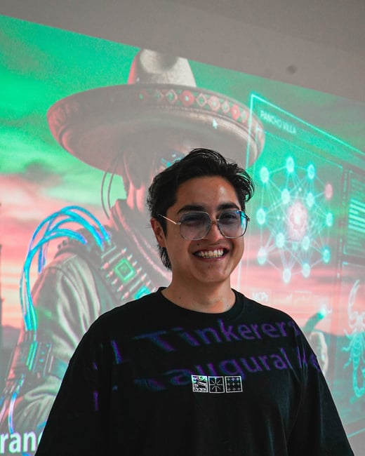 A young man with glasses and a black t-shirt smiles in front of a projected image of a futuristic character wearing a sombrero.