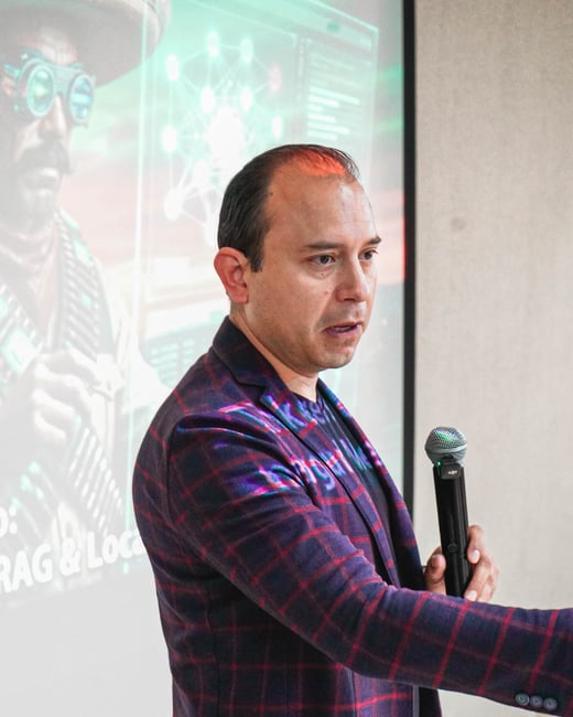 A man in a purple and red checkered blazer is giving a presentation, holding a microphone in front of a projector screen.