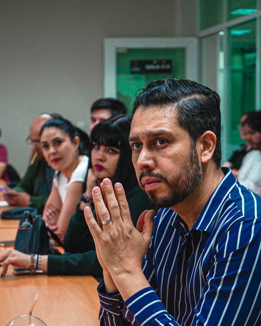 A man in a blue striped shirt sits at a wooden table, looking attentively off-camera with his hands clasped. Behind him, several other people are also seated, appearing to be part of a meeting or presentation.
