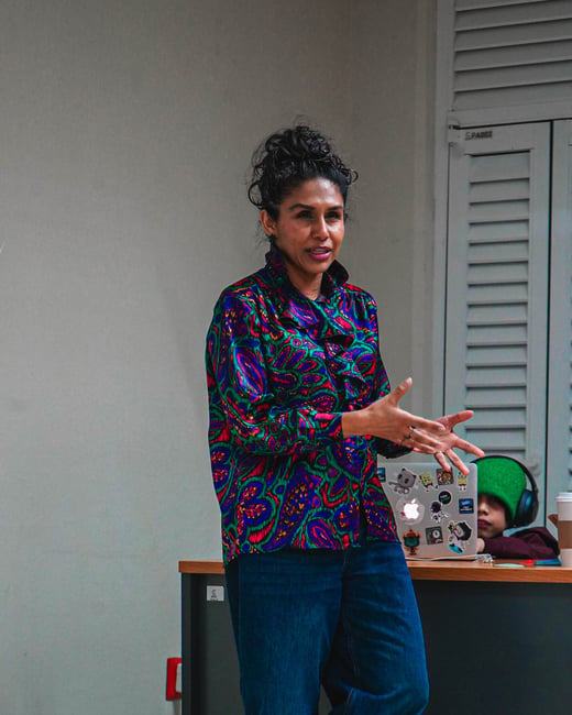 A woman in a vibrant, patterned blouse and blue jeans stands gesturing while speaking in an indoor setting, with a second person visible behind a sticker-covered laptop.