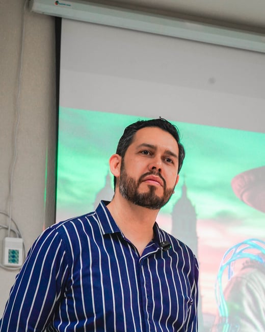 A man with a beard wearing a blue and white striped shirt stands in front of a projection screen during a presentation.