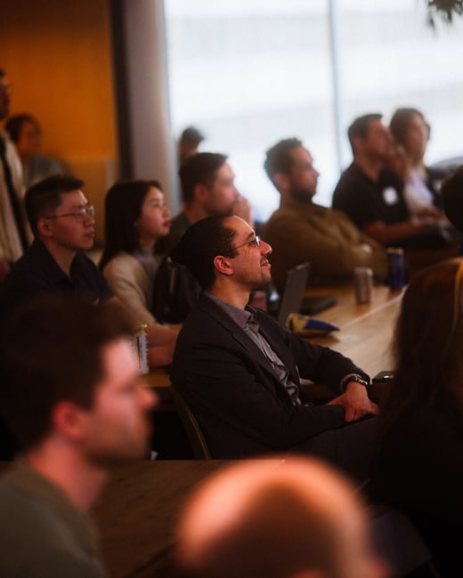 A group of people sitting at tables in an indoor setting, appearing to listen to a presentation or speaker off-camera.