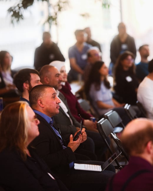 A group of people sitting in an audience, attentively listening to a presentation or speaker off-camera in an indoor setting.