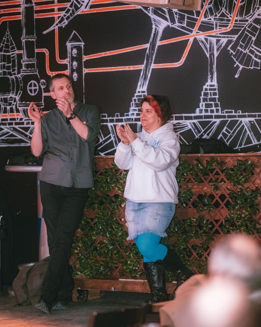 A man and a woman are standing in front of a stylized industrial mural, clapping for an audience.