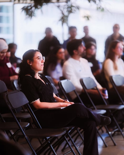 A woman with glasses and dark hair sits in a folding chair, attentively watching a presentation alongside other audience members in an indoor setting.