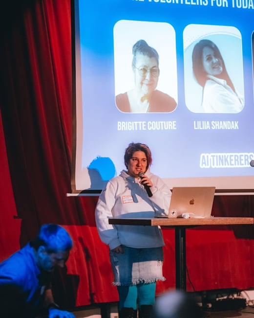 A person stands on a stage giving a presentation, with a projection screen behind them displaying names and photos of volunteers.