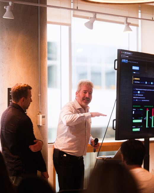 A man stands in an indoor office setting, gesturing toward a large digital display while holding a microphone during a presentation.