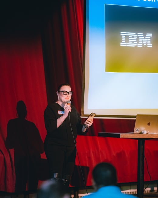 A woman stands on a stage, speaking into a microphone and holding a smartphone during a presentation, with an IBM logo projected on a screen behind her.