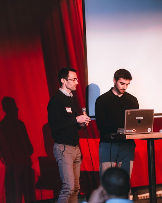 Two men are on a stage in front of a red curtain; one holds a microphone while the other operates a laptop on a high table.