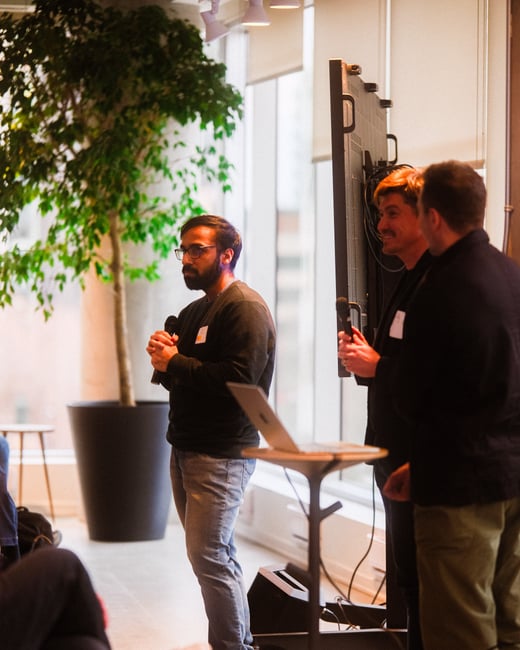 A man with a beard and glasses is speaking into a microphone during a presentation in a modern indoor space with large windows and a potted tree.