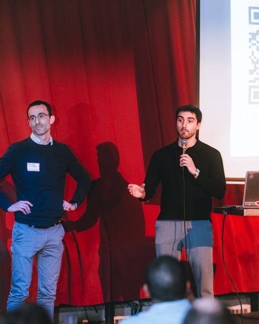Two men are giving a presentation on a stage with a red curtain background. One man is speaking into a microphone while the other stands next to him with his hands on his hips.