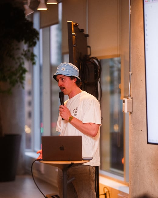 A man wearing a light blue bucket hat and a white t-shirt is speaking into a microphone while standing behind a laptop at a presentation.