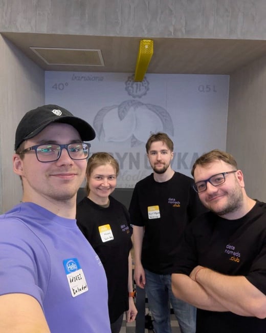 A group selfie featuring four people (three men and one woman) indoors, likely at a professional or networking event, all wearing name tags and three wearing black shirts branded with "data nomads .club".