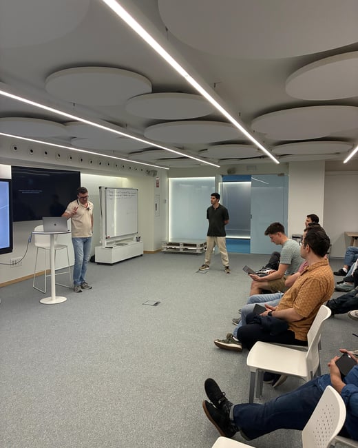 A man delivers a presentation to a small group of people in a modern office or event space characterized by distinctive circular ceiling panels.