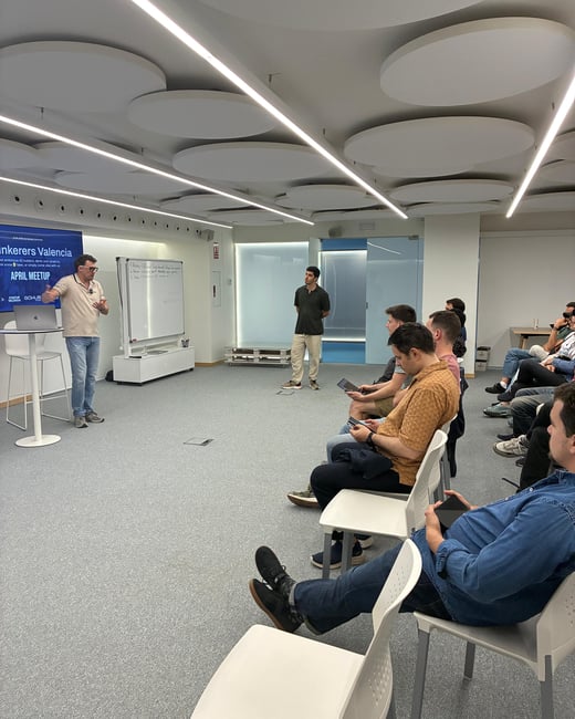 A man stands at the front of a modern room giving a presentation to a seated audience during a 'Tinkerers Valencia' meetup.