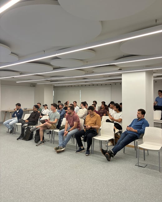 A group of people are seated in an audience-style arrangement within a modern, minimalist room, likely attending a presentation or talk.
