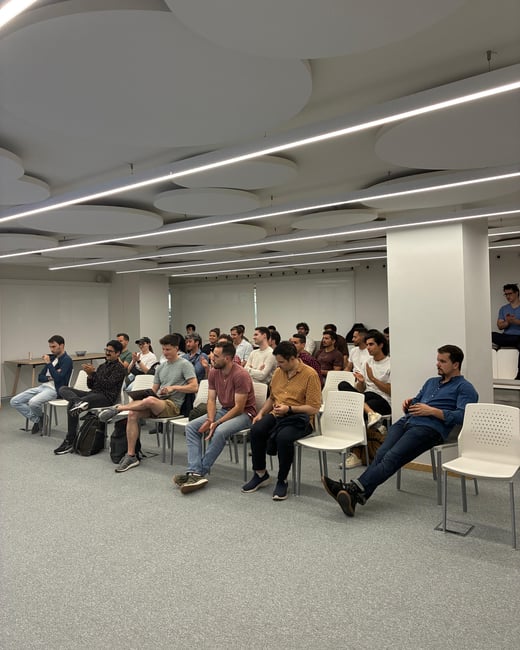A group of people, primarily young men, are seated in white chairs in a modern indoor space, appearing to be an audience for a presentation or meeting.