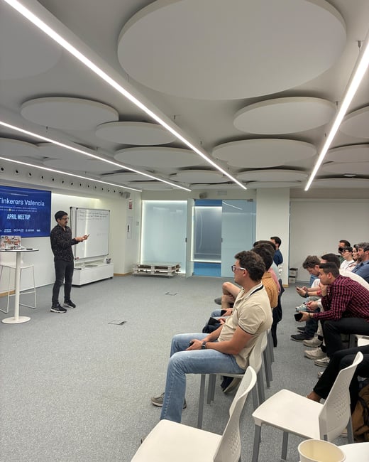 A man is giving a presentation to a seated audience in a modern indoor space with distinctive circular ceiling panels.