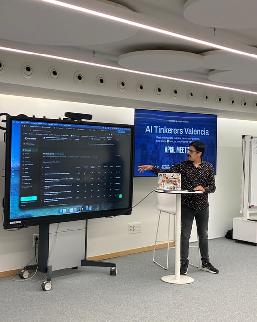 A man is giving a presentation in a modern office setting, pointing at a large interactive display showing a data dashboard while a secondary screen displays event branding for 'AI Tinkerers Valencia'.