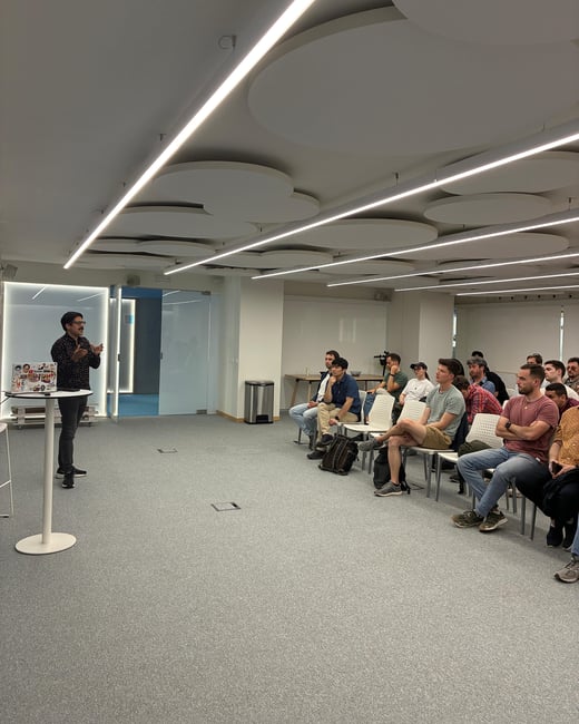 A man is giving a presentation to a seated audience in a modern office or event space featuring distinctive circular ceiling panels and linear lighting.