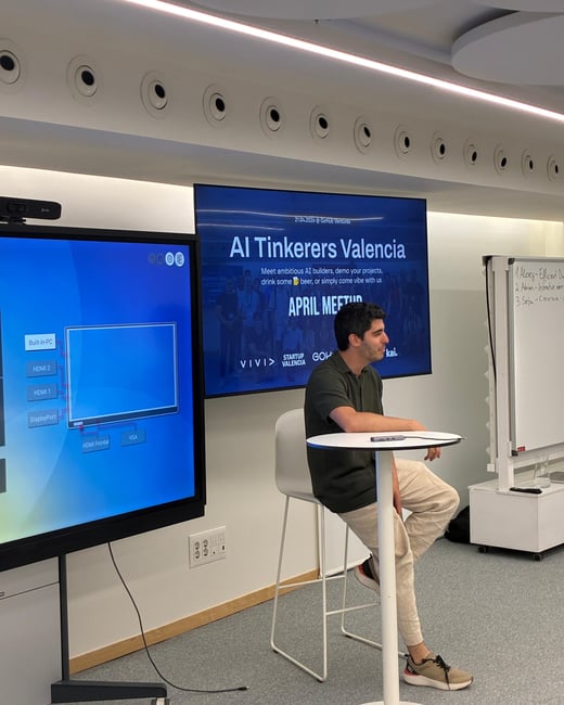 A man sits on a high stool in a modern event space, positioned between a large touchscreen displaying input options and a screen showing a presentation for 'AI Tinkerers Valencia'.