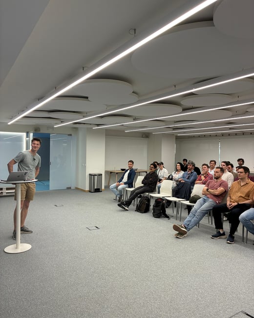 A man stands at a tall table with a laptop, delivering a presentation to a seated audience in a modern indoor space with circular ceiling panels.