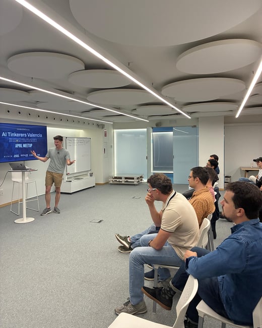 A man stands in front of a digital screen giving a presentation titled 'AI Tinkerers Valencia' to a small audience in a modern indoor space.