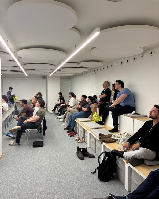 A group of people are seated in a modern, minimalist room with tiered bench seating and circular ceiling panels, likely attending a presentation or workshop.
