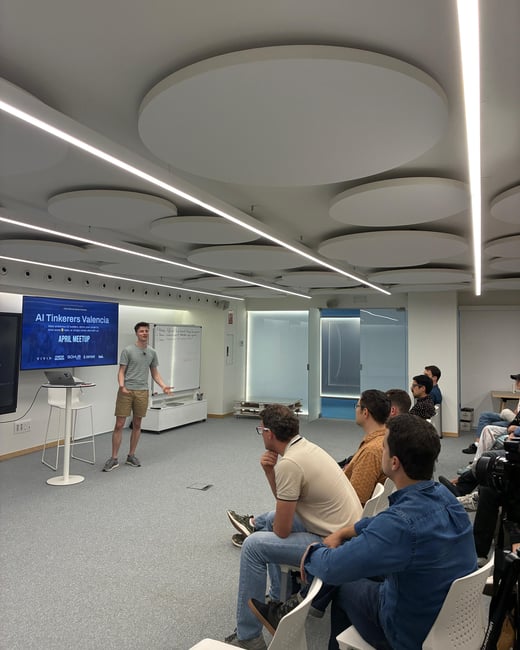 A young man is giving a presentation to a seated audience in a modern, brightly lit room with circular ceiling panels.
